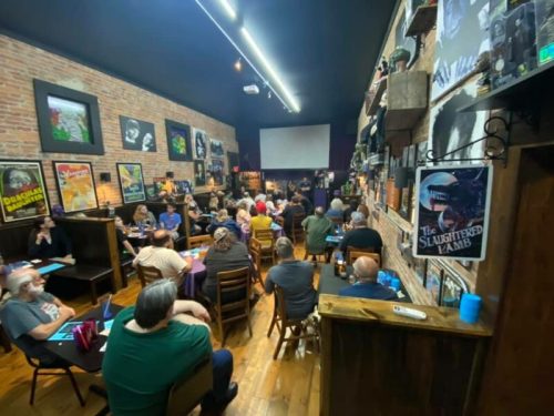 A crowded room with people seated at tables, watching a presentation on a stage with movie posters on the walls.