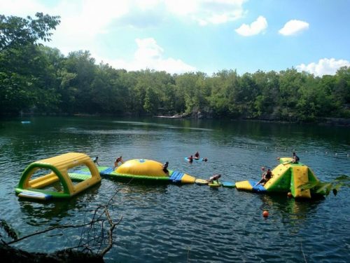 Colorful inflatable water park on a lake, with people playing and swimming in the background surrounded by trees.