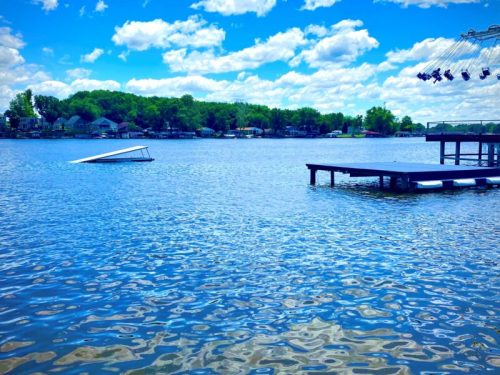 A serene lake scene with a dock, a tilted boat, and fluffy clouds reflecting in the water under a bright blue sky.