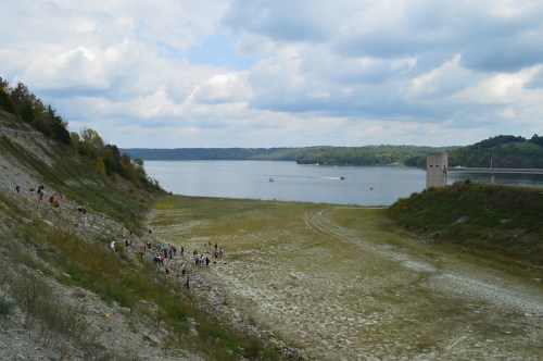 A dry lakebed with people exploring, surrounded by hills and a distant dam under a cloudy sky.