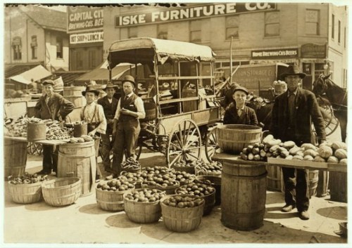 A historic market scene with vendors selling produce from barrels and carts, set in a bustling town square.