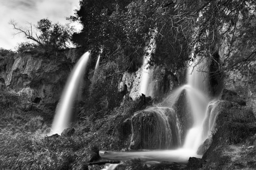 A serene black and white image of cascading waterfalls surrounded by lush greenery and rocky terrain.