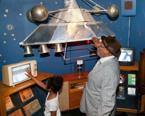 An adult and a child interact with a retro display featuring a spaceship model and vintage electronics in a museum.