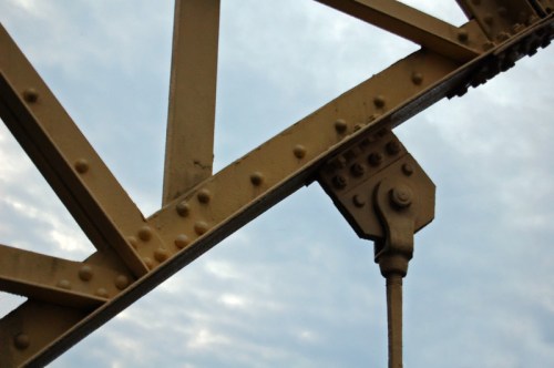 Close-up of a yellow metal bridge structure with rivets against a cloudy sky.