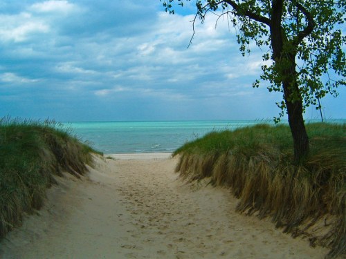 A sandy path between grassy dunes leads to a calm, turquoise sea under a cloudy sky.