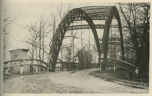 Black and white photo of an arched metal bridge over a dirt road, surrounded by bare trees.