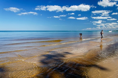 Two people walking along a sandy beach with calm waters and a clear blue sky dotted with clouds.