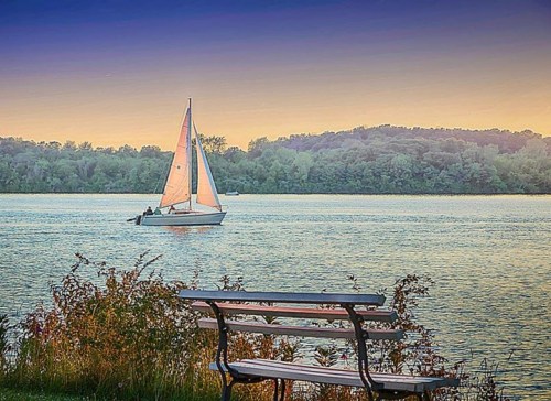 A serene lake scene at sunset with a sailboat gliding by and a bench in the foreground.