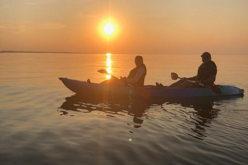 Two people kayaking on calm water at sunset, with the sun reflecting on the surface.