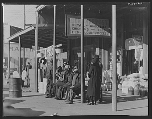 A group of people in long coats sitting outside a storefront, with signs for groceries and feed in the background.