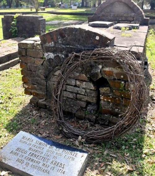 A weathered grave with a stone marker and a circular vine wreath in a cemetery, surrounded by greenery.