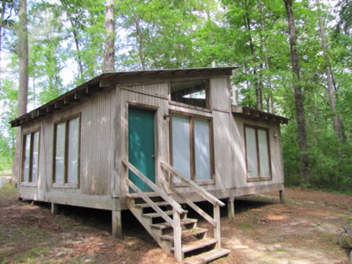 A wooden cabin with green doors and large windows, surrounded by trees in a forested area.