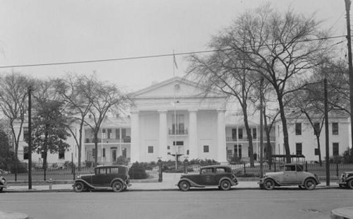 Historic black-and-white photo of a large white building with columns, surrounded by trees and vintage cars parked in front.
