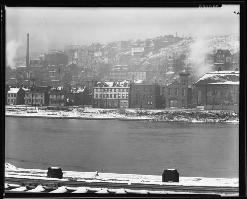 Black and white photo of a snowy riverside town with industrial buildings and a hillside in the background.