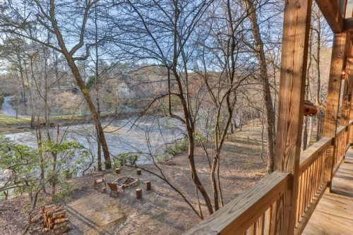 View from a wooden balcony overlooking a serene, wooded area with a fire pit and a winding river in the distance.