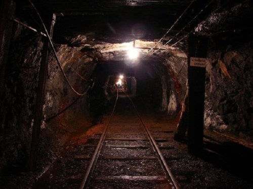 Dimly lit underground mine tunnel with railway tracks and rocky walls.