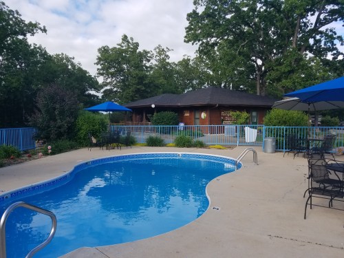 A serene pool area with blue water, surrounded by greenery and patio furniture, under a partly cloudy sky.