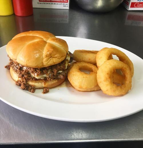 A plate with a sloppy joe sandwich on a bun and three crispy onion rings, set against a kitchen background.