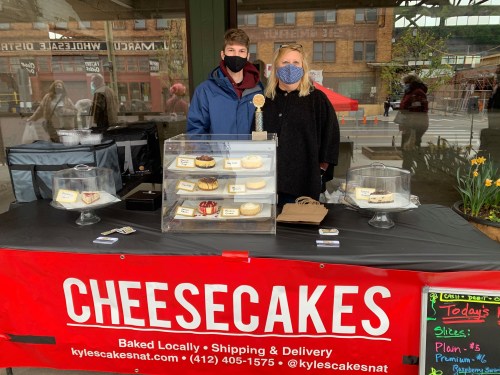 A man and woman stand behind a display of cheesecakes at a market booth, wearing masks and smiling.