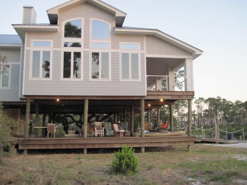 A modern house on stilts with large windows, surrounded by trees and a deck with outdoor seating.