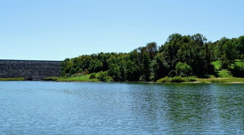 A serene lake surrounded by lush greenery and a dam in the background under a clear blue sky.