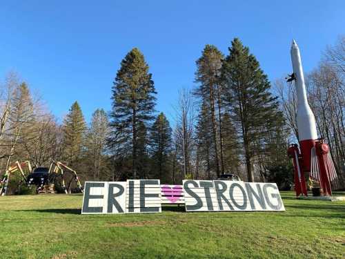 A large sign reading "ERIE ❤️ STRONG" in a grassy area, with trees and a rocket sculpture in the background.