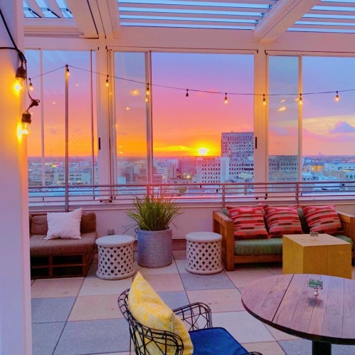 A rooftop lounge with cozy seating and string lights, overlooking a vibrant sunset and city skyline.