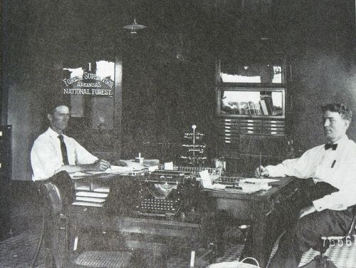 Two men seated at a desk in an office, surrounded by typewriters and paperwork, with a sign for the National Forest.