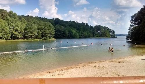 A serene lake surrounded by trees, with people swimming and a sandy beach in the foreground under a blue sky.