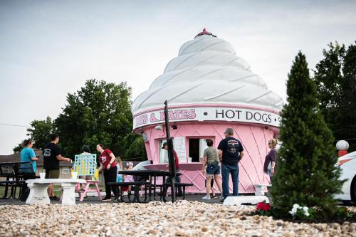 A pink ice cream-shaped stand with people ordering food, surrounded by trees and gravel.