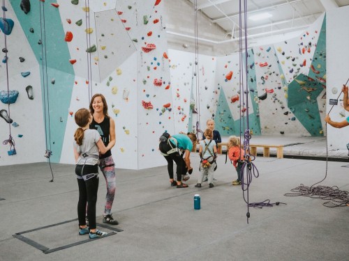 A group of children and adults at an indoor climbing gym, preparing for climbing activities on colorful walls.