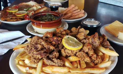 A plate of fried chicken with lemon slices and fries, surrounded by various side dishes and drinks on a table.