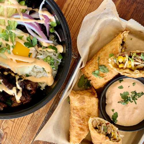 A bowl of rice topped with vegetables and sauce, alongside crispy fried rolls and a small bowl of dipping sauce.