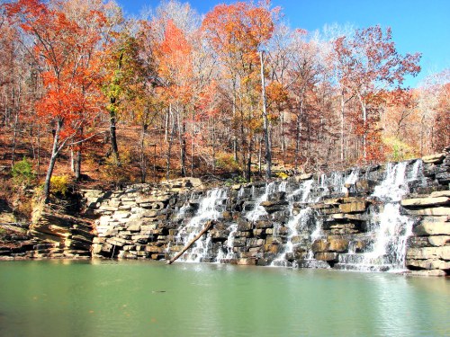 A serene waterfall cascades over rocks, surrounded by vibrant autumn foliage and a clear blue sky.