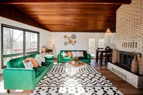 Cozy living room with green sofas, a patterned rug, and a stone fireplace, featuring wooden beams and large windows.