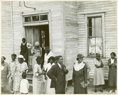 A group of people, dressed in early 20th-century attire, gathered outside a wooden building.