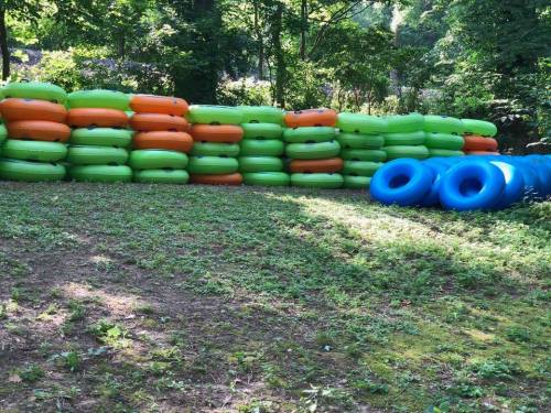 A stack of colorful inflatable tubes in green, orange, and blue, arranged on a grassy area surrounded by trees.