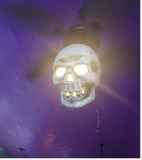 A ceiling fan with a skull-shaped light fixture, glowing from within, against a purple ceiling.