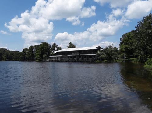 A serene lake scene featuring a wooden building surrounded by trees under a blue sky with fluffy clouds.