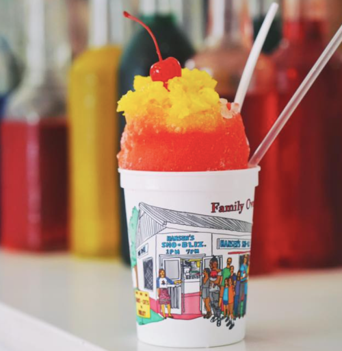 A colorful snow cone in a cup with a cherry on top, set against a backdrop of vibrant syrup bottles.