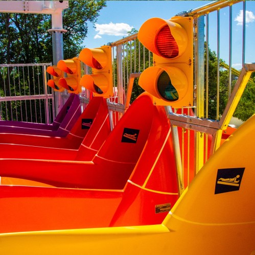 Colorful seats in orange and purple line up under traffic lights at an amusement park ride.
