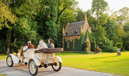A vintage car with a bride and groom drives past a charming stone chapel surrounded by lush greenery.