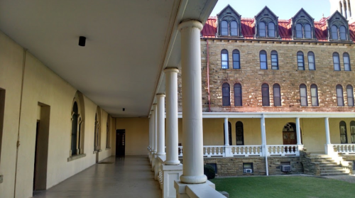A corridor with columns leading to a historic stone building with arched windows and a red roof.