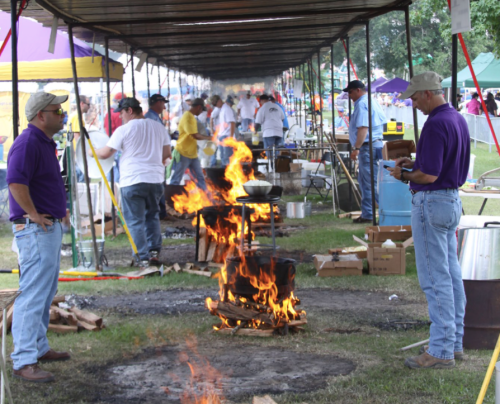 Two men in purple shirts stand near a fire pit at an outdoor cooking event, surrounded by tents and people.