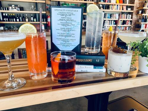 A wooden bar with a variety of colorful cocktails and a menu, surrounded by books on a shelf in the background.