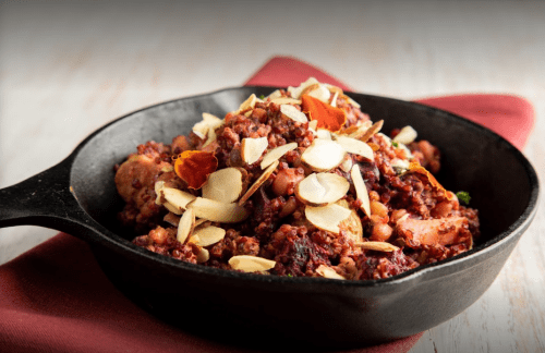 A cast iron skillet filled with a colorful grain dish topped with sliced almonds and garnished with herbs.