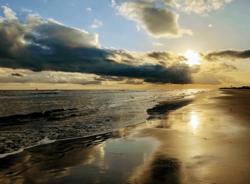 A serene beach at sunset, with golden reflections on wet sand and dramatic clouds in the sky.