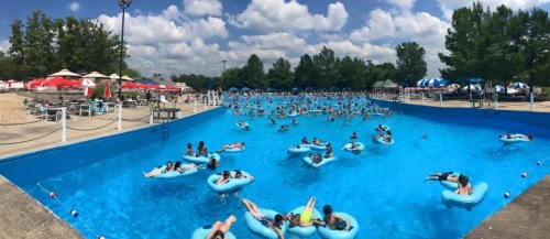 A crowded water park with people floating on inner tubes in a large blue pool under a sunny sky.