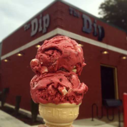 A hand holding a cone with two scoops of red ice cream in front of a brick building labeled "The Dip."