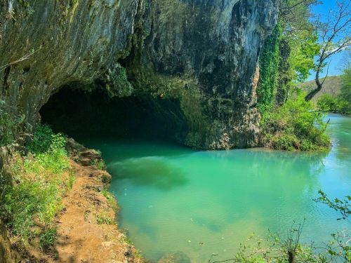 A serene cave entrance by a turquoise river, surrounded by lush greenery and rocky cliffs.
